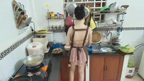 Video of an Asian woman in a kitchen, wearing a pink apron and black hair tied up, cooking at a stove with a black countertop, surrounded by various kitchen utensils and appliances.