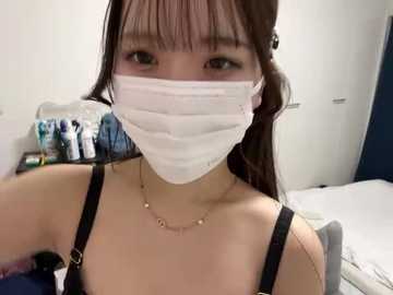 Video of an East Asian woman with long brown hair, wearing a white surgical mask and black bra, seated in a medical room.