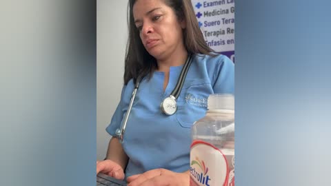 Video of a Hispanic nurse with medium skin tone, wearing a light blue scrub top and stethoscope, looking at a tablet, holding a water bottle in a medical office with a wall sign.