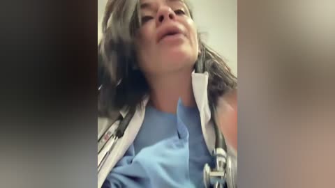 Video of a woman in a blue shirt, wearing a white lab coat with a stethoscope around her neck, looking intently at something.