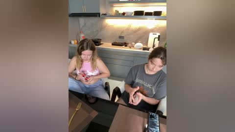 Video of two women in a modern kitchen, one wearing a pink top, the other a grey t-shirt, both sitting at a table, focused on their phones.