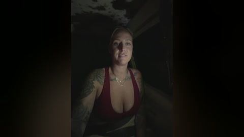 Video of a blonde woman with tattoos, wearing a red low-cut top, standing in a dimly lit, shadowy tunnel.