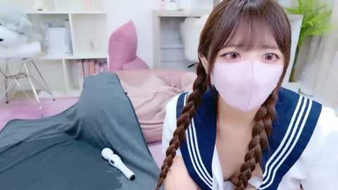 A video of a young East Asian woman with long brown pigtails, wearing a Japanese schoolgirl uniform and a pink face mask, lying on a bed in a soft-pink room.