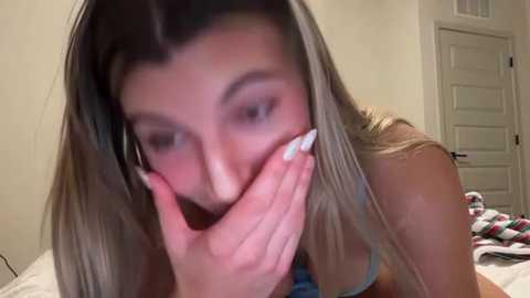Video of a young woman with long, straight, blonde hair, wearing a light blue bra, covering her mouth with her hands in a bedroom with a white door and a bed with a plaid blanket.