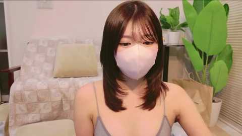 Video of an East Asian woman with straight, shoulder-length dark brown hair, wearing a white mask, grey spaghetti strap top, in a modern living room with beige furniture, green plant, and blinds.