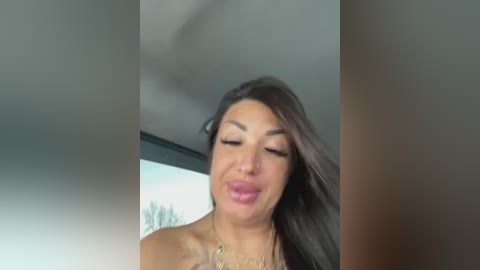 Video of a smiling, light-skinned woman with long brown hair, wearing a gold necklace, indoors by a window with a blurred outdoor view.