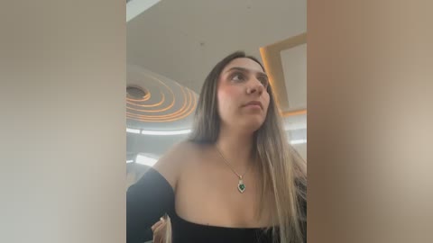 A video of a young woman with long, straight brown hair, wearing a black off-the-shoulder top, standing indoors against a modern, circular ceiling with warm lighting.