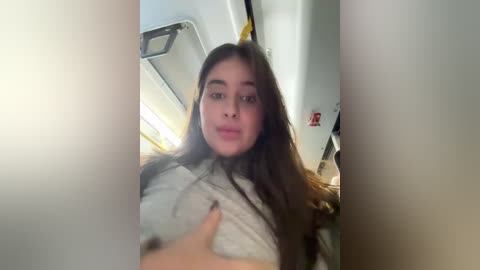 Video of a young woman with long brown hair, wearing a grey sweater, inside an airplane cabin, with a hand on her breast.