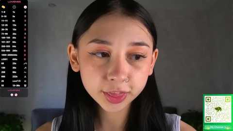 Video of an Asian woman with long black hair, fair skin, and subtle makeup, wearing a nose ring, in a modern indoor setting.