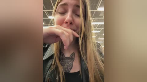 Video of a young woman with long, blonde hair, wearing a black leather jacket, with a large wolf tattoo on her chest, looking introspective, in a fluorescent-lit room.