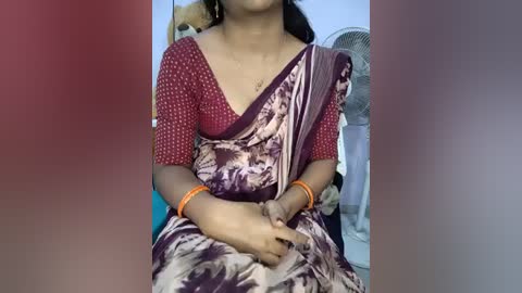 Video of a woman in a maroon and white sari with a polka-dot blouse, wearing an orange bracelet. She sits on a bed with a fan and clothes in the background.