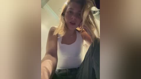 A video of a young Caucasian woman with blonde hair, wearing a tight white tank top and blue jeans, captured from a slightly blurry, angled perspective.