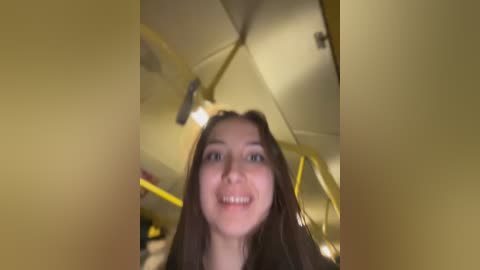 Video of a young woman with long brown hair and fair skin, smiling, standing inside a yellow umbrella, with blurred background suggesting a bright, possibly sunny setting.