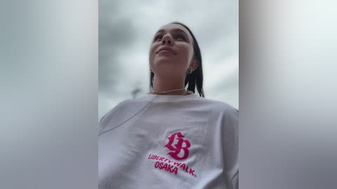 Video of a young woman with straight, dark hair, wearing a white t-shirt with a red logo and text in Cyrillic script, standing against a blurred, overcast sky.