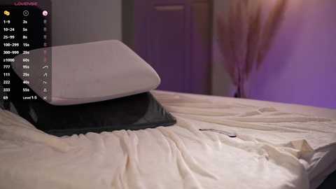 Video of a minimalist bedroom with a neatly made bed featuring a white pillow atop a black pillow on a beige bedspread. A smartphone and remote control rest on the bed. The background is a blurred, softly lit wall with purple hues.