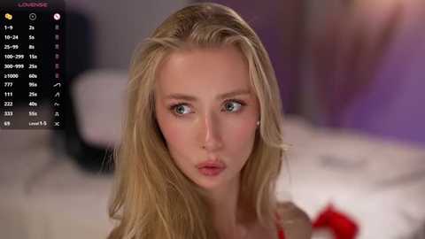 Video of a blonde woman with fair skin, blue eyes, and long hair, looking serious and thoughtful. Background shows a blurred bedroom with a bed and purple walls.
