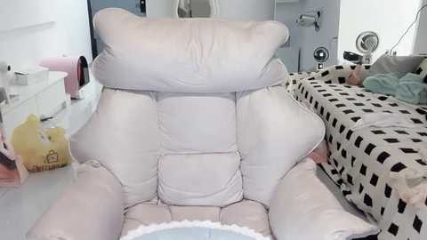 Video of a messy, cream-colored recliner chair in a small, cluttered living room with a white bed adorned with black and white patterned sheets, a white dresser, and a laundry basket.