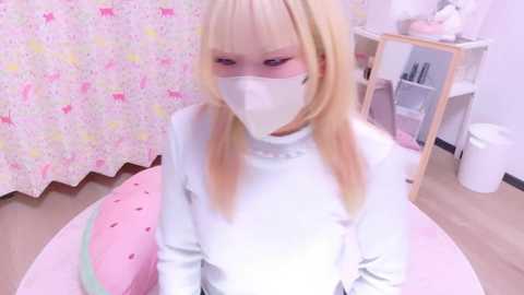 Video of a young, pale-skinned, blonde-haired girl in a white turtleneck and face mask, sitting on a pink, watermelon-shaped cushion in a room with a pink, cartoon-patterned curtain and a white trash can.