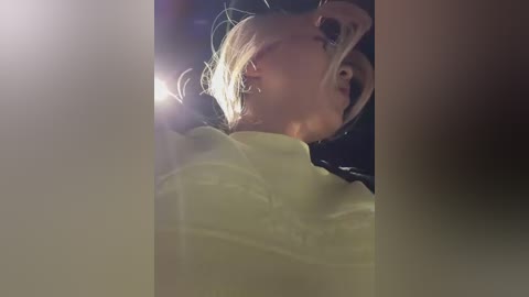 A video captures a young blonde girl with light skin, wearing a yellow sweater, looking up with a smile. The background is blurry and dark, with lens flares.