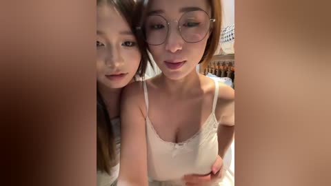 Video of two young Asian women with light skin, one with glasses, wearing white camisoles, posing closely together, blurred background.