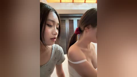 A video of two young Asian women, one with long black hair, wearing a grey top, and the other with brown hair in a red scrunchie, sitting side by side.