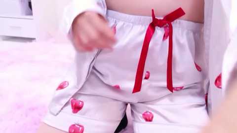 Video of a person wearing white pajamas with red cherries, a red ribbon tie, and a white robe, sitting on a pink fluffy carpet.