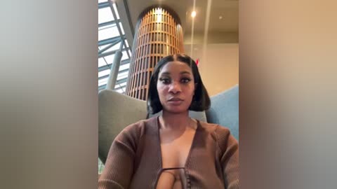 A video of a young woman with medium brown skin and short, straight black hair, wearing a brown cardigan over a low-cut top, standing in a modern, brightly lit room with large windows and a cylindrical wooden lamp.