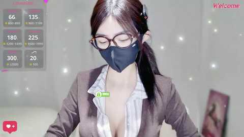Video of an Asian woman with long black hair, wearing a black face mask, glasses, and a brown blazer over a white shirt with a cleavage reveal. Background is blurred with a \"Welcome\" text.