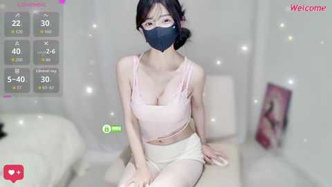 Video of an East Asian woman with fair skin, wearing a pink tank top, black face mask, and white shorts, sitting on a bed in a softly lit room.
