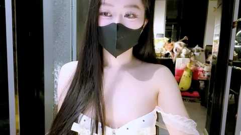 Video of an East Asian woman with long black hair, fair skin, and a black face mask, wearing a white, off-the-shoulder dress. Background shows a cluttered room with colorful objects and a window.