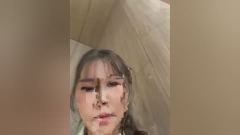 Video of a young woman with medium skin tone and long, dark hair, partially obscured by a reflective, distorted glass surface. The background is neutral, possibly a wall, with soft lighting.