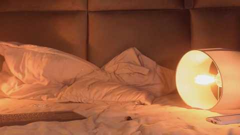 Video of a cozy bedroom with a lit bedside lamp casting a warm glow on rumpled white sheets and pillows, set against a beige, padded headboard.
