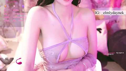 Video of an East Asian woman with light skin, wearing a revealing, strappy lavender lace top, exposing ample cleavage, sitting on a bed with pink and purple hues.