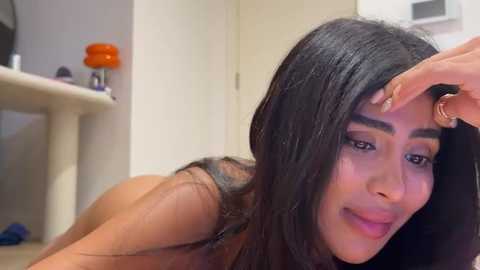 Video of a young woman with long black hair, light brown skin, and dark eyes, lying on her stomach, smiling, with a hand gently touching her forehead, in a modern, minimalistic room with a beige wall and a shelf with an orange pumpkin and a few small items.