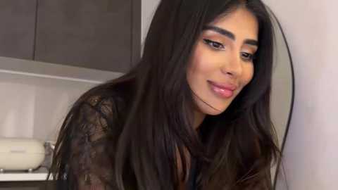 Video of a Middle Eastern woman with long, straight black hair, wearing a black lace top, in a modern kitchen with grey cabinets and a white backsplash.
