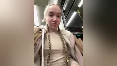 Video of a young woman with long, blonde braided hair, wearing a beige sweater and brown jacket, sitting on a public bus.