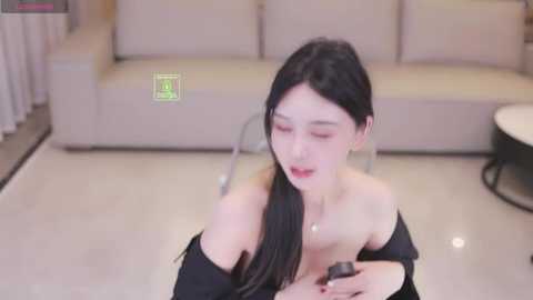Video of an East Asian woman with long black hair, wearing a black off-shoulder top, sitting on a light-colored floor, holding a phone, in a modern living room with beige sofa and white curtains.