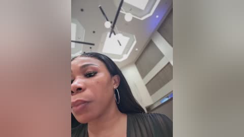 Video of a dark-skinned Black woman with long straight black hair, wearing hoop earrings, making a pouty face. She is indoors with a modern, minimalist ceiling with recessed lighting and large windows.