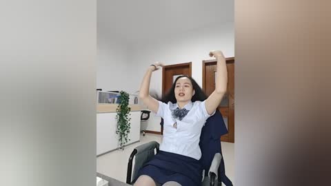 Video of a joyful Asian woman with straight black hair, wearing a white blouse and navy skirt, raising her arms in an office with a desk, potted plant, and wooden door.