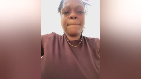 Video of a middle-aged Black woman with medium-brown skin, wearing a maroon shirt, and a gold necklace. Her hair is styled in braids. The background is a blurred, light-colored room.