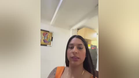 Video of a Latina woman with long, dark hair, medium skin tone, and a tattoo on her arm, wearing an orange top, standing in a modern kitchen with a colorful painting on the wall and a fridge in the background.