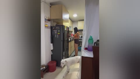 A video captures a woman in an orange sports bra and black shorts, standing in a modern kitchen. A white dog stands beside her. The kitchen features stainless steel appliances and a countertop cluttered with items.