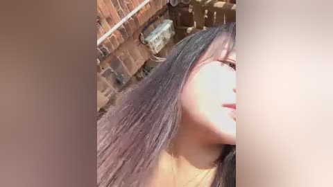 Video of a young woman with long, straight, dark hair, partially obscured by shadow, in a brick alleyway. She has light skin and is wearing a light-colored top.