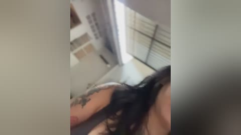 A blurry video captures a tattooed arm with a black flower design, partially obscured by a blurred figure in the background. The setting appears to be an indoor room with white walls and a window with closed blinds.