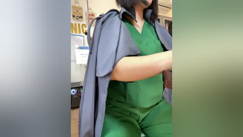 Video of a person with light brown skin and dark hair, wearing green scrubs and a grey lab coat, standing in a brightly lit medical office with posters on the wall.