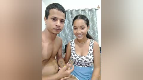 Video of a shirtless man with a small penis, and a smiling woman in a patterned top, standing in a room with gray curtains.