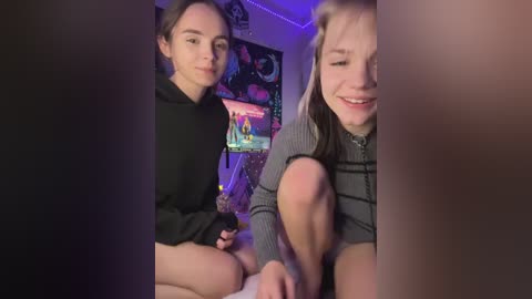 Video of two young people, a smiling woman with light hair and a boy with brown hair, sitting closely, both wearing casual clothes, in a dimly lit room with colorful posters and a purple light strip.