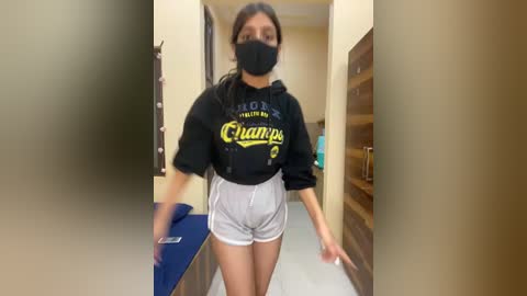 Video of a young woman with medium skin tone, wearing a black face mask, black hoodie, and gray shorts, walking down a beige hallway.
