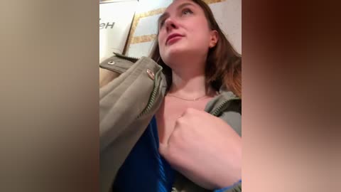 Video of a young woman with fair skin, brown hair, and blue top, seen from a low angle, holding a green jacket, in a dimly lit room with blurred background.