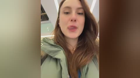 Video of a young Caucasian woman with long brown hair, wearing a green jacket, sticking out her tongue in a playful manner. Background shows a blurred indoor setting with beige and green tones.